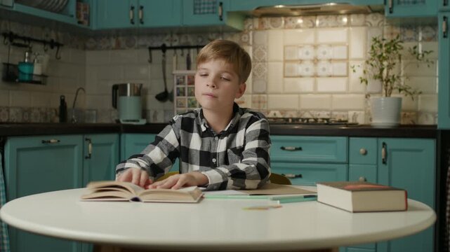 School boy studying at home, writing in a notebook while checking an open book for homework.