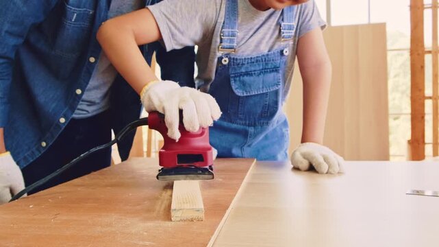 Two people wearing gloves sand a wooden board with a red electric sander on a workbench, practicing safe DIY woodworking in a bright home workshop.