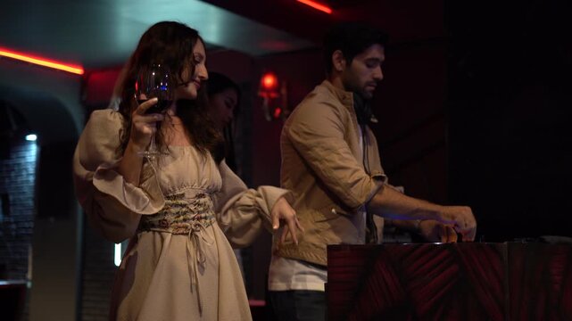 Happy woman friends dancing, toasting, and holding wine glasses in a nightclub. Man DJ is mixing music in the background. Young people having fun and celebrating. Nightlife.