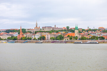 Obraz premium Budapest historic Buda side unfolds along the Danube River under overcast sky. Skyline features the spires of Matthias Church and Fisherman's Bastion rising above colorful riverside buildings