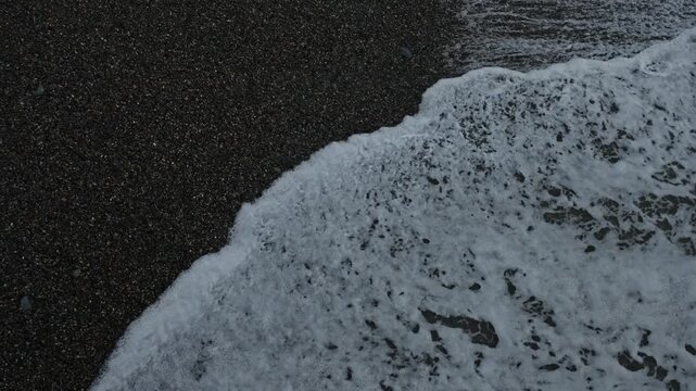 Black pebble beach with foamy surf and textured backwash, closeup aerial perspective of dark volcanic sand, rhythmic waves drawing