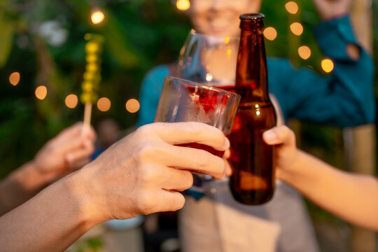 Friends clinking beer bottle, wine glass, and cocktail at outdoor party with warm bokeh string lights, shallow depth of field, celebrating together.