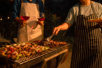 Two people in aprons grilling kebabs and steak on outdoor barbecue at night, holding red wine glasses, warm lighting, casual dinner party atmosphere. © Thitaporn