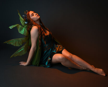 portrait of female model wearing fantasy fairy costume, green dress, cloak, butterfly wings and crown. full length sitting pose, kneeling on floor, dark studio background.