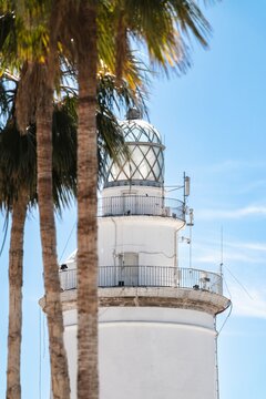 White lighthouse in Malaga framed by palm trees under a clear blue sky. Coastal landmark, navigation tower and Mediterranean travel destination.