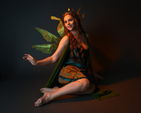 portrait of female model wearing fantasy fairy costume, green dress, cloak, butterfly wings and crown. full length sitting pose, kneeling on floor, dark studio background.
