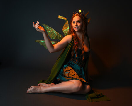 portrait of female model wearing fantasy fairy costume, green dress, cloak, butterfly wings and crown. full length sitting pose, kneeling on floor, dark studio background.