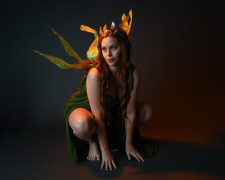 portrait of female model wearing fantasy fairy costume, green dress, cloak, butterfly wings and crown. full length sitting pose, kneeling on floor, dark studio background.