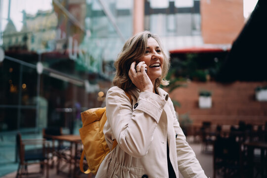Portrait of happy middle aged Caucasian woman with yellow backpack phoning in city, cheerful female traveler using smartphone for roaming call near cafe, positive vibes, authentic representation.