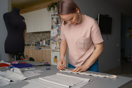 Woman seamstress measuring decorative trim at table in a home sewing studio, focused on garment making