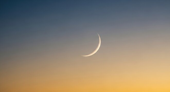 Minimalist view of a thin crescent moon in a clear evening sky featuring a smooth color gradient transitioning from dark blue at the top to warm orange and yellow at the horizon line.