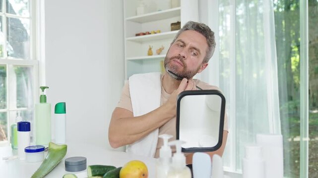 Bearded man combing beard in morning light. Bearded man grooming beard at home spa. Middle aged man combing beard for daily hygiene. Bearded man shaping beard in front of mirror.