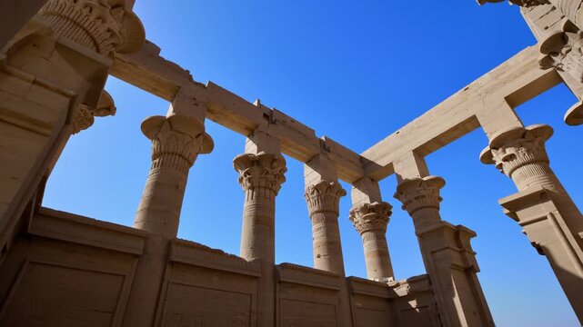 Looking up through carved sandstone columns with decorative papyrus capitals and stone crossbeams at Philae temple, Aswan, Egypt, clear blue sky above.
