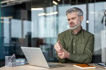 Obraz premium Senior man working on a laptop at an office desk, gripping his aching wrist due to discomfort, showing symptoms of carpal tunnel syndrome or workplace injury from overuse