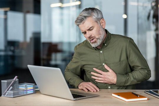 Mature man having severe chest pain at his office desk. Holding his chest with discomfort and looking distressed while sitting in front of a laptop. Indicating a serious health problem or heart attack