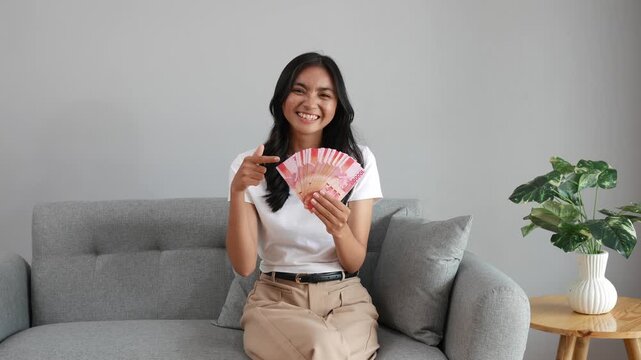 Cheerful Asian woman holding Indonesian rupiah banknotes while sitting on couch at home room