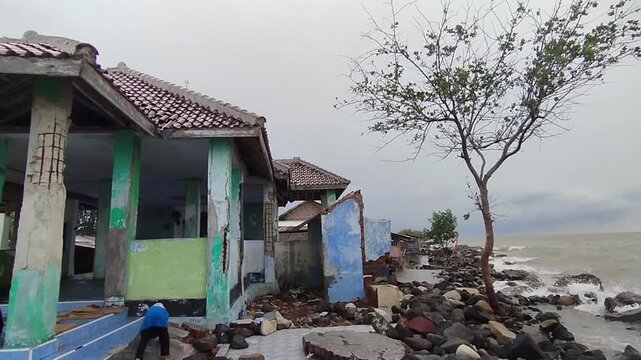 DEMAK, INDONESIA - JANUARY 4, 2025: Abandoned house being destroyed by coastal erosion and sea level rise on a rocky beach under a cloudy sky.