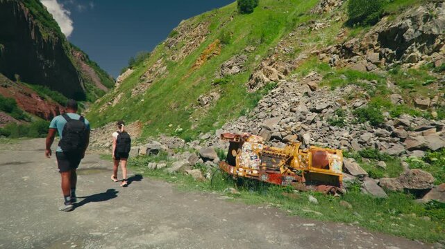 A couple walks along a gravel path in the mountains, passing an old abandoned bulldozer. Rustic industrial relic meets scenic nature in a unique outdoor adventure setting.