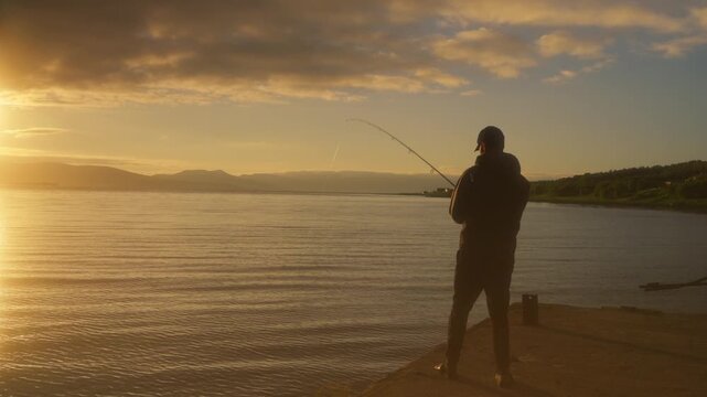 A shoreline fisherman gets a bite on his line at sunset, with golden light shimmering across the fjord and the sun setting behind distant mountains in Norway.