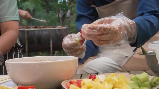 Close-up of gloved hands preparing fresh fruit salad outdoors at a picnic table, with bowl and chopped pineapple, apple and strawberry in foreground, casual cooking scene.