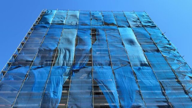 Blue construction safety net on building scaffolding moving in wind. Urban renovation process and dynamic fabric motion on facade structure concept.