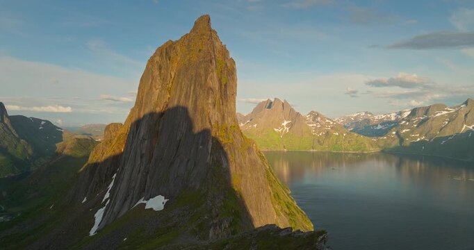 Drone flies toward Segla mountain on Senja island, Norway. People stand on a rock admiring fjord views, with calm water reflecting surrounding snow-capped peaks.
