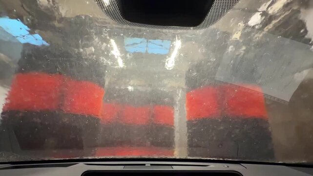 View from inside car during automatic car wash process with rotating brushes and water jets cleaning windshield. Interior perspective through wet glass with foam, reflections and blurred lights.