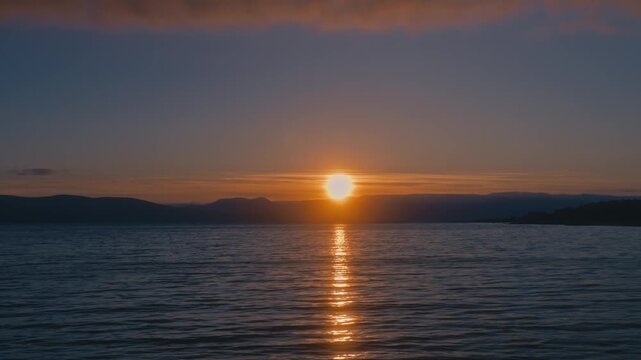 Golden midnight sun over Arctic ocean waters with distant mountains on the horizon in Northern Norway. Warm light reflects on calm sea, blending sunset and sunrise tones.