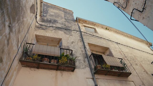 An upward view of weathered walls and modest balconies with flower pots captures the lived-in texture, warm light, and timeless charm of a Mediterranean village home.