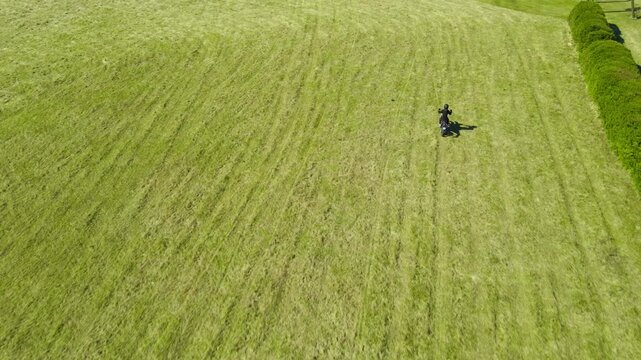 Aerial high angle view showing a lone motorcyclist riding across a mown grass field, eventually reaching and following a dirt road through the sunny, rural landscape in springtime
