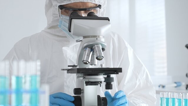 Male doctor in protective suit and gloves using microscope while analyzing samples while conducting research in sterile laboratory. Close-up of test tubes with blue liquid