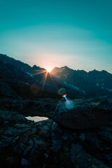 Obraz premium Intense sunburst with dramatic lens flare over dark alpine mountain silhouette, deep blue sky and rocky terrain at sunrise, Italian Alps