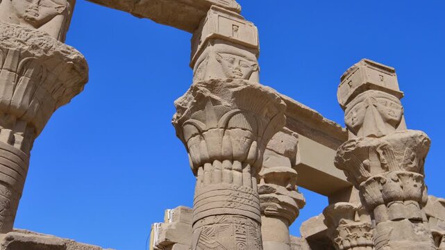 Dramatic upward perspective of carved Hathor goddess columns at Philae temple, Aswan, Egypt, stone lintels with hieroglyphic inscriptions crossing vivid blue sky.