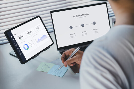 Remote work planning scene with professional writing strategy notes beside laptop and tablet displaying business dashboard analytics, data-driven workflow, modern workspace productivity concept
