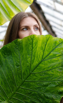 Woman holding large leaf in greenhouse showing her eyes and part of her face in nature setting