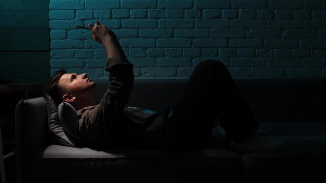 Young man lying on a sofa at night, scrolling through content on his smartphone, the blue light from the screen illuminating his face in a dark room with brick wall