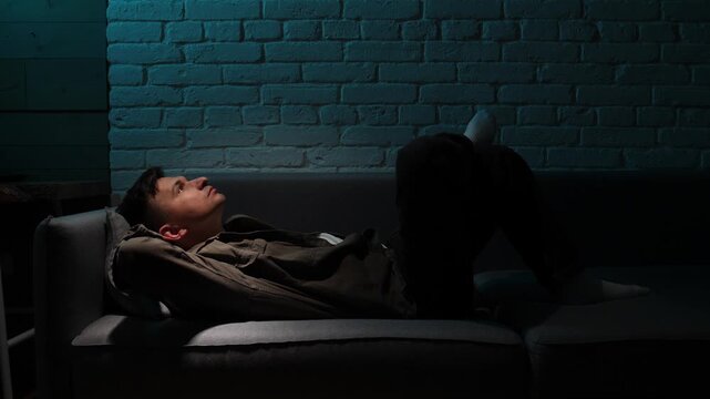 Man lying awake on a sofa in a dark living room, sleepless and pensive, wrestling with insomnia, loneliness and heavy thoughts late at night