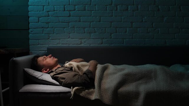 Young man feeling sad and stressed, lying awake on a couch at night, unable to sleep due to anxiety, grief, and suffering from a sleep disorder in a dark room