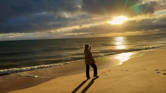 Woman holding mobile phone in hand and taking sunrise photo. Woman using smart phone take a photo at evening on the beach of sunset. Travel photo, copy space.