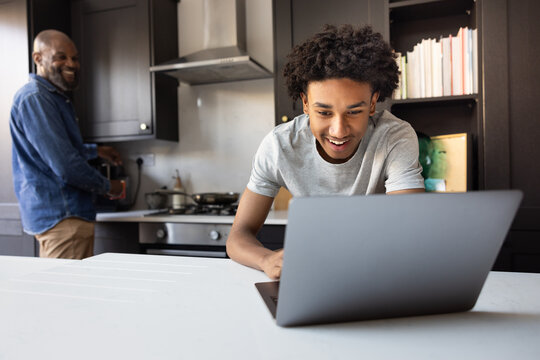 Teenager using laptop while father prepares coffee