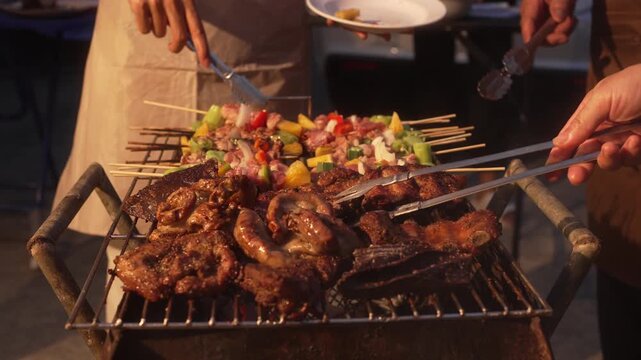 Close-up of outdoor barbecue grill with steaks, sausages and colorful vegetable skewers being turned by people at a summer cookout, warm evening light.