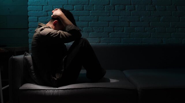 Young man sitting on a dark sofa with his head in his hands, lonely and anxious, struggling with depression and emotional distress in a moody, low key interior