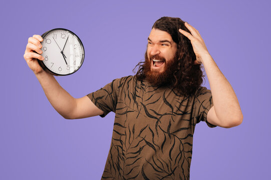 Bearded White Man Holding Clock, Surprised Expression, Purple Studio Backdrop, Casual Tshirt, Hand Scratching