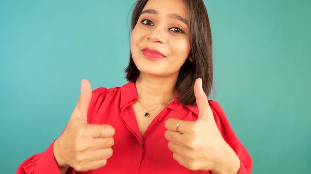 Close-up of cheerful young Asian woman girl showing thumbs up sign gives positive feedback look at camera nodding head wears red shirt posing against blue studio background.