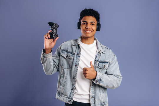 Young male gamer wearing headphones holds a video game controller while giving a thumbs up, standing against a solid purple background