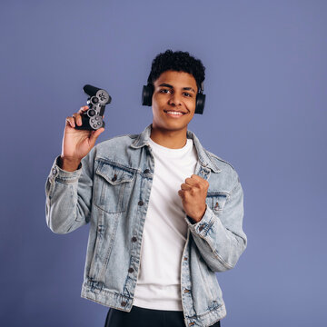 Young male gamer wearing headphones holds a video game controller while smiling against a solid purple background, showcasing a modern gaming lifestyle and technology