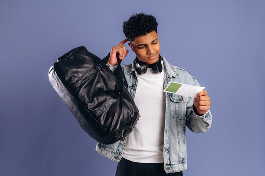 Young African American male student holding a travel itinerary while carrying a black duffel bag, wearing headphones and a denim jacket against a purple background