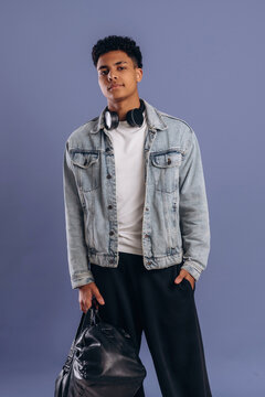 Young male model wearing a denim jacket and headphones poses confidently with a black bag against a solid purple background in a studio setting