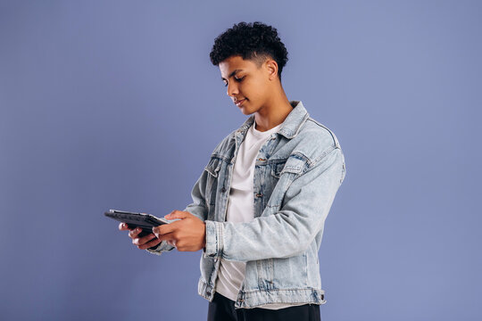 Young male holding a tablet while wearing a denim jacket and white t-shirt, standing against a solid light purple background