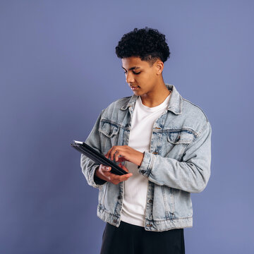 Young male student wearing a denim jacket and white t-shirt holds a tablet while standing against a solid purple background in a studio setting